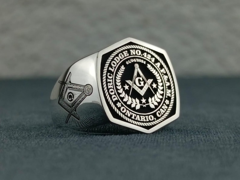 Puede incluir: Un anillo mas&oacute;nico de plata con un dise&ntilde;o grabado en blanco y negro. El anillo presenta una escuadra y un comp&aacute;s, una 'G' y el texto "Doric Lodge No. 424 A.F.&A.M. Ontario, Can."