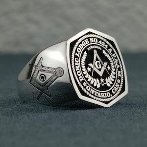 Puede incluir: Anillo mas&oacute;nico de plata con un dise&ntilde;o de esmalte negro. El anillo presenta una br&uacute;jula y una escuadra con una 'G' en el centro, rodeados de una corona y el texto "Doric Lodge No. 424 A.F. & A.M. Ontario, Can."