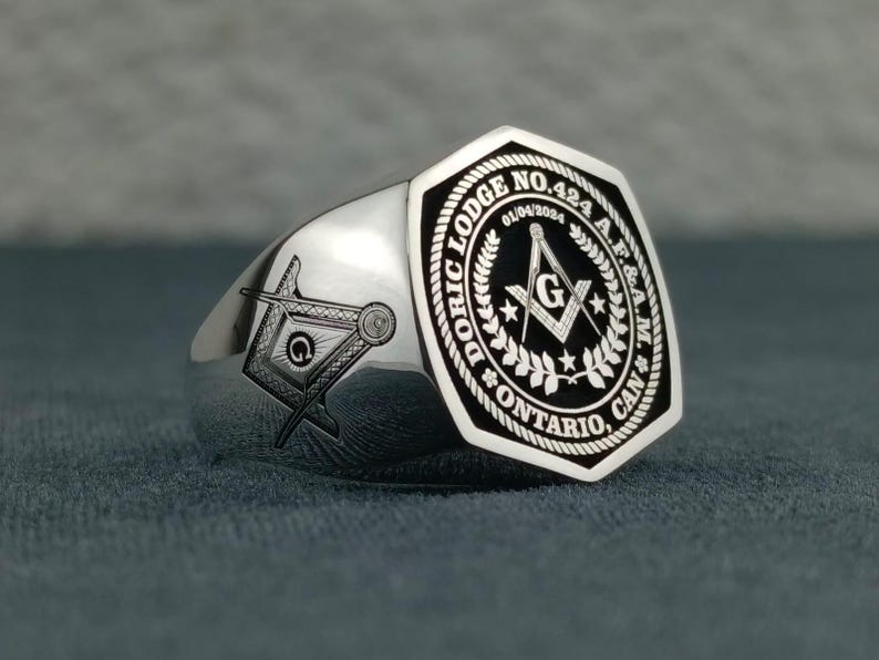 Puede incluir: Anillo mas&oacute;nico de plata con un dise&ntilde;o de esmalte negro. El anillo presenta una br&uacute;jula y una escuadra con una 'G' en el centro, rodeados de una corona y el texto "Doric Lodge No. 424 A.F. & A.M. Ontario, Can."