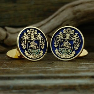 May include: A pair of round gold-toned cufflinks featuring a deep blue enamel background. Each cufflink displays a detailed gold crest with a mounted knight, ornate scrollwork, and a banner at the bottom with the text SHUBIANOK in gold lettering.