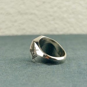 Puede incluir: Un anillo de plata con un dise&ntilde;o de cuadrado y comp&aacute;s grabado en el lateral. El anillo est&aacute; hecho de un metal liso y pulido y tiene una forma redondeada.