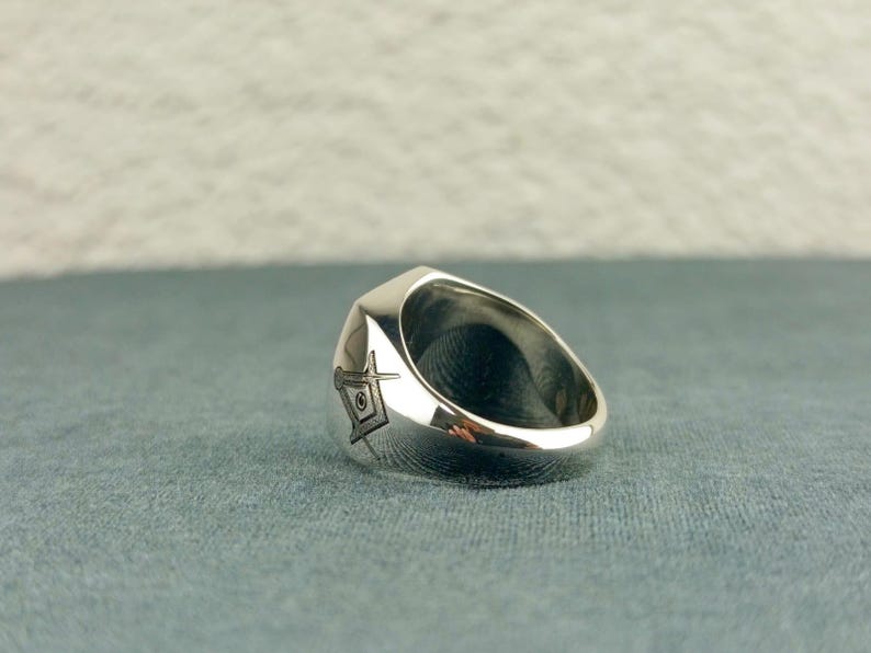 Puede incluir: Un anillo de plata con un dise&ntilde;o de cuadrado y comp&aacute;s grabado en el lateral. El anillo est&aacute; hecho de un metal liso y pulido y tiene una forma redondeada.