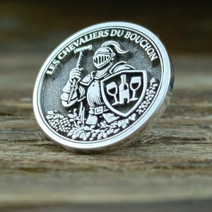May include: Silver ring with the text "LES CHEVALIERS DU BOUCHON" around the edge. The ring features a knight holding a hammer, a shield with a bottle and two glasses, and grape vines.