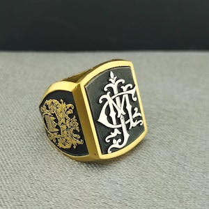 Könnte beinhalten: Ein schwarzer und goldener Siegelring mit einem weißen Monogramm und einem Wappen-Design. Der Ring ist aus Metall und hat eine polierte Oberfläche.