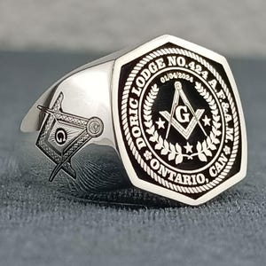 Puede incluir: Un anillo mas&oacute;nico de plata con un dise&ntilde;o grabado en blanco y negro. El anillo presenta una escuadra y un comp&aacute;s, una estrella y el texto "DORIC LODGE NO. 424 A.F.&A.M. ONTARIO, CAN."