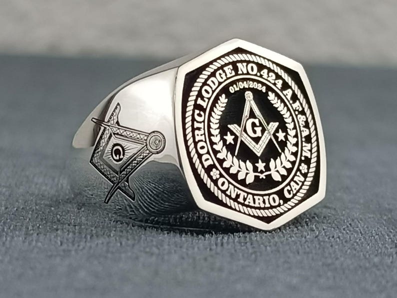 Puede incluir: Un anillo mas&oacute;nico de plata con un dise&ntilde;o grabado en blanco y negro. El anillo presenta una escuadra y un comp&aacute;s, una estrella y el texto "DORIC LODGE NO. 424 A.F.&A.M. ONTARIO, CAN."
