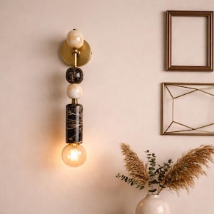 May include: A modern wall sconce with a brass finish and a decorative design of stacked marble and stone elements. The sconce features a warm, glowing light bulb. The background includes framed artwork and a decorative vase.