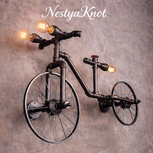Puede incluir: Una lámpara de pared con forma de bicicleta hecha de tuberías de metal negro. La lámpara tiene tres bombillas en el manillar y una en el asiento. Las ruedas también son de metal. El texto "Nestya Knot" está en la parte superior.