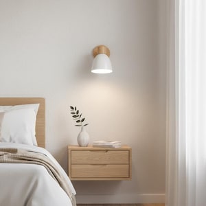 Puede incluir: Escena de dormitorio moderno con una lámpara de pared blanca con base de madera, que ilumina una mesita de noche flotante de madera. La mesita de noche tiene dos cajones, un pequeño jarrón blanco con vegetación y un libro. Se ve una cama con ropa de cama blanca y una manta beige.