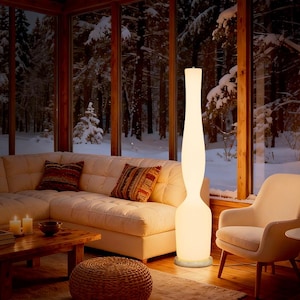May include: A tall, cream-colored floor lamp with a curved design stands in a cozy living room. The lamp is illuminated, casting a warm glow. The room features a white sectional sofa, patterned throw pillows, and a wooden coffee table. A snowy forest is visible through the large windows.
