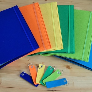 Könnte beinhalten: Ein Satz von sieben Filzordnern in Regenbogenfarben, blau, orange, gelb, grün, lindgrün und hellblau, mit einem blauen Filzband an jedem Ordner. Die Ordner sind in einem Halbkreis auf einer Holzoberfläche angeordnet. Es gibt sechs bunte Filzschlüsselanhänger in den gleichen Farben wie die Ordner.