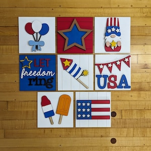 May include: Eight wooden signs with a patriotic theme. The signs feature a star, balloons, a gnome, fireworks, a banner, a popsicle, an ice cream cone, and the words "Let freedom ring" and "USA".