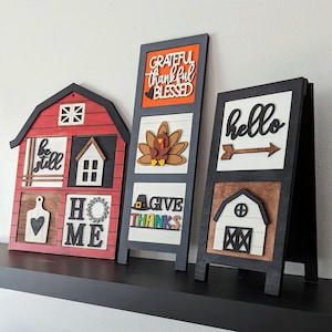 May include: Three wooden signs with a fall theme. The first sign is red and says "be still, home". The second sign is black and says "grateful thankful blessed, give thanks". The third sign is black and says "hello".