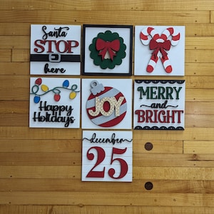 May include: Six wooden Christmas signs with various holiday themes. The signs feature red, green, white, and black paint. The signs include "Santa Stop Here", "Happy Holidays", "Joy", "Merry and Bright", "December 25", and a wreath with a red bow.