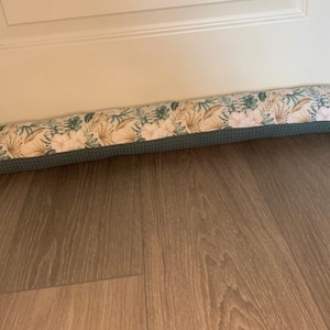 Könnte beinhalten: Ein langer, rechteckiger Türstopper mit einem Blumenmuster in Grün-, Weiß- und Beigetönen. Der Türstopper ist aus Stoff und hat eine graue Rückseite.