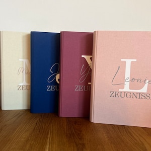 Könnte beinhalten: Fünf verschiedene farbige, stoffbezogene Ordner mit dem Wort "ZEUGNISSE" und einem großen Anfangsbuchstaben auf der Vorderseite. Die Ordner sind in einer Reihe von links nach rechts angeordnet, wobei die Farben grau, beige, dunkelblau, burgunderrot und rosa sind.