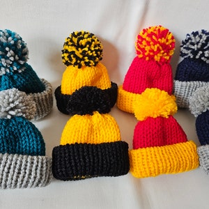 May include: Five knitted hats with pom-poms. The hats are in various colours, including teal, yellow, red, and black. The pom-poms are also in various colours, including white, black, and yellow.