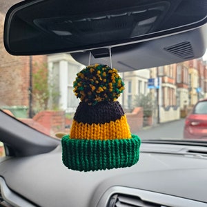 May include: A green, black and yellow knitted hat with a pom-pom hangs from the rear view mirror of a car.