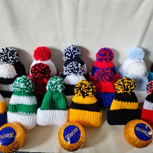 May include: A collection of ten knitted hats with pom-poms in various colors and patterns, including black and white, red and blue, green and white, and yellow and black. The hats are arranged in a row on a white surface, with two chocolate candies in the foreground.