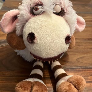 May include: A plush toy cow with a white and cream-colored body, pink ears, and brown horns. The toy has a stitched mouth with visible teeth and blood-red accents around the eyes and nose. The legs are striped brown and white, with brown hooves.