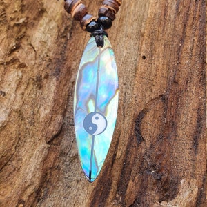 Könnte beinhalten: Eine schwarze Schnurkette mit braunen und schwarzen Holzperlen und einem Surfbrett-Anhänger aus schillerndem Abalone-Muschel mit einem Yin-Yang-Symbol.