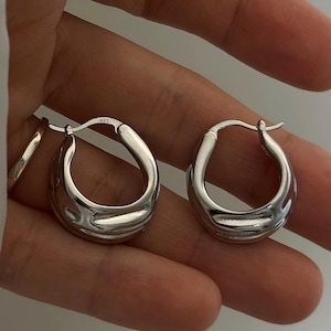 以下が含まれることがあります： テクスチャ加工された有機的なデザインの、銀色のフープピアス2つ。ピアスは手で持たれており、磨かれた反射面が際立っています。フープは、モダンで大胆な外観をしています。