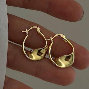 Pode incluir: Um par de brincos de argola dourados com um design assimétrico único. Os brincos têm um acabamento liso e polido e uma forma subtil e orgânica. Os brincos são segurados numa mão.