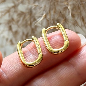 Puede incluir: Un par de pendientes de aro dorados con forma rectangular. Los pendientes son pequeños y delicados, y tienen un diseño simple y moderno.