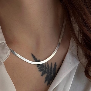 Op de afbeelding: Een zilveren visgraatketting. De ketting is plat en ligt soepel op de huid. De achtergrond omvat een crèmekleurige blouse.