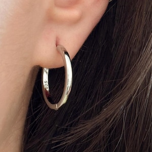 Puede incluir: Pendientes de aro de plata con un acabado liso y pulido. Los pendientes se llevan en la oreja de una persona.