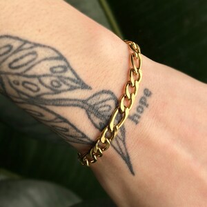 Puede incluir: Un brazalete de cadena dorada con un diseño de eslabones de curb. El brazalete está en la muñeca de una persona.