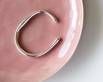 Puede incluir: Un brazalete de plata abierto con un diseño curvo y liso. El brazalete está apoyado en un plato de cerámica rosa.