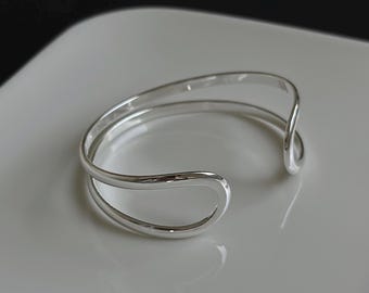 May include: A silver bangle bracelet with a simple, modern design. The bracelet is made of two curved bands that intersect, creating a unique and stylish look.