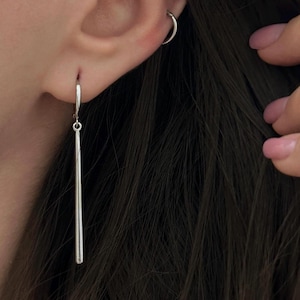 May include: A silver bar earring hangs from a silver hoop earring. The earring is a long, slender, polished silver bar. A small silver hoop earring is also visible in the ear. Dark hair is in the background.