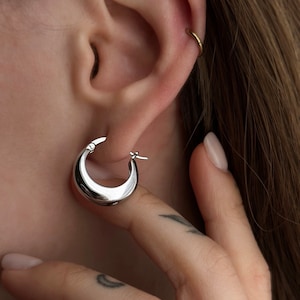 May include: Silver hoop earrings with a curved, rounded design. The earrings are worn in the earlobe.