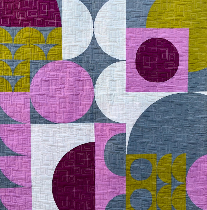 May include: A quilt with a geometric pattern in shades of pink, purple, yellow, and gray. The quilt is made up of circles, squares, and half-circles.
