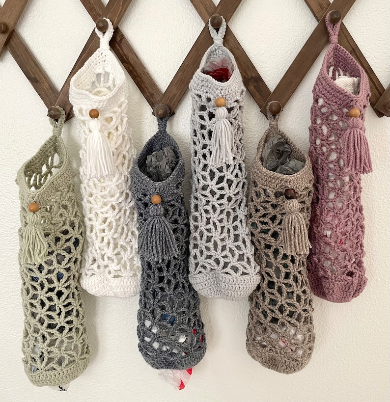 May include: Six crocheted bags hanging on a wooden rack. The bags are in various colors, including beige, gray, white, and pink. Each bag has a tassel hanging from the bottom.
