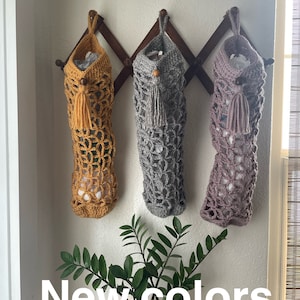 May include: Three crocheted bags hanging on a wooden rack. The bags are in mustard yellow, gray, and light purple. The bags have a tassel at the bottom. The text "New colors" is written below the bags.