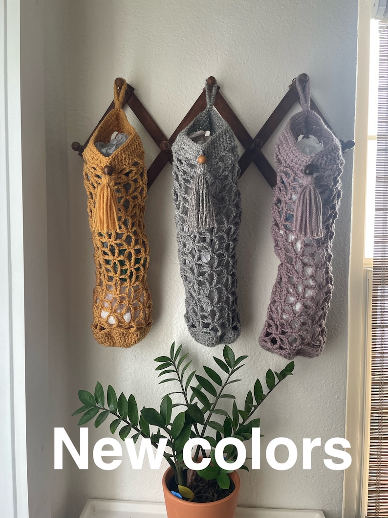 May include: Three crocheted bags hanging on a wooden rack. The bags are in mustard yellow, gray, and light purple. The bags have a tassel at the bottom. The text "New colors" is written below the bags.