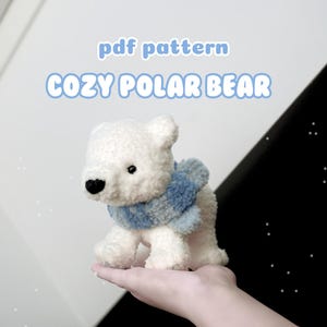 Könnte beinhalten: Ein weißer gehäkelter Eisbär, der einen blau-weißen Schal trägt. Der Bär sitzt in der Hand einer Person. Der Text "pdf pattern cozy polar bear" befindet sich über dem Bären.