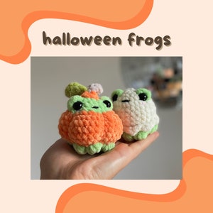 May include: Two crocheted frog plushies. One is orange and shaped like a pumpkin with green accents. The other is white with green accents. The text "halloween frogs" is above the plushies.