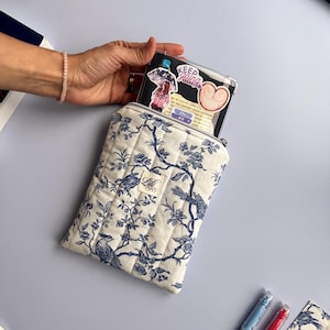 Peut inclure: Une housse de téléphone matelassée crème avec un motif floral et d'oiseaux bleu. L'étui est ouvert, révélant une coque de téléphone transparente avec des autocollants. L'étui a une fermeture éclair et une petite étiquette qui dit "ARS".