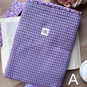 Op de afbeelding: Een lavendelkleurige boekhoes van wafelstof met een bloemenpatroon en bijpassende bladwijzer. De hoes heeft een klein wit label met de tekst "Lili ARS". Een boek is gedeeltelijk zichtbaar, samen met een kaart met "have a spl".