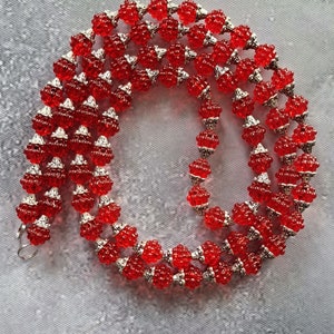 Op de afbeelding: Een rode kralenketting met zilverkleurige accenten. De ketting heeft kleine, ronde, rode kralen gescheiden door zilverkleurige metalen kralen. De ketting wordt gepresenteerd op een grijze ondergrond.