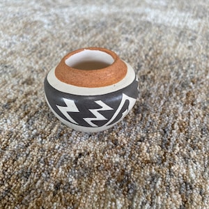 May include: A small, round ceramic pot with a wide opening. The pot features a design with a white base, a dark gray band with a zigzag pattern, and a brown rim. The interior is white.