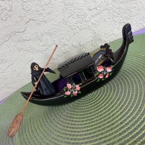 May include: A black ceramic gondola with a skeletal figure rowing. The gondola is decorated with pink roses and a small coffin with a cross on top.