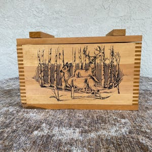 May include: A wooden box with a hinged lid and a decorative deer scene. The box is made of light-colored wood with a darker wood trim. The front of the box features a detailed illustration of deer in a forest setting.