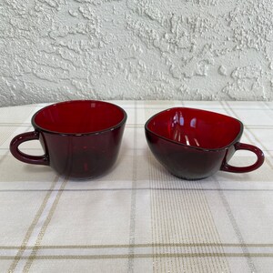 May include: Two red glass cups, one with a rounded shape and a handle, the other with a square shape and a handle. The cups are on a white and beige checkered tablecloth.