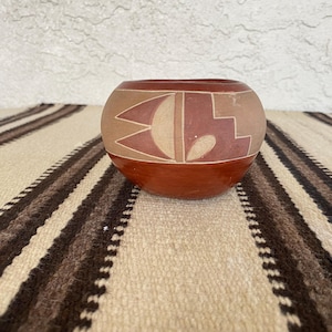 May include: A round, hand-painted ceramic bowl with a reddish-brown base and a tan upper section. The bowl features geometric designs in shades of tan and reddish-brown. The bowl is sitting on a striped textile.