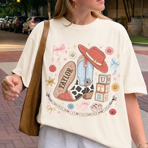 Pode incluir: Uma t-shirt cor de creme com uma impress&atilde;o gr&aacute;fica de uma bota de cowboy com padr&atilde;o de vaca, um chap&eacute;u de cowboy vermelho, blocos de alfabeto com as letras UTS, uma estrela de xerife, la&ccedil;os, flores e o texto TAYLOR e I KNEW IT YOU KNEW YOU.